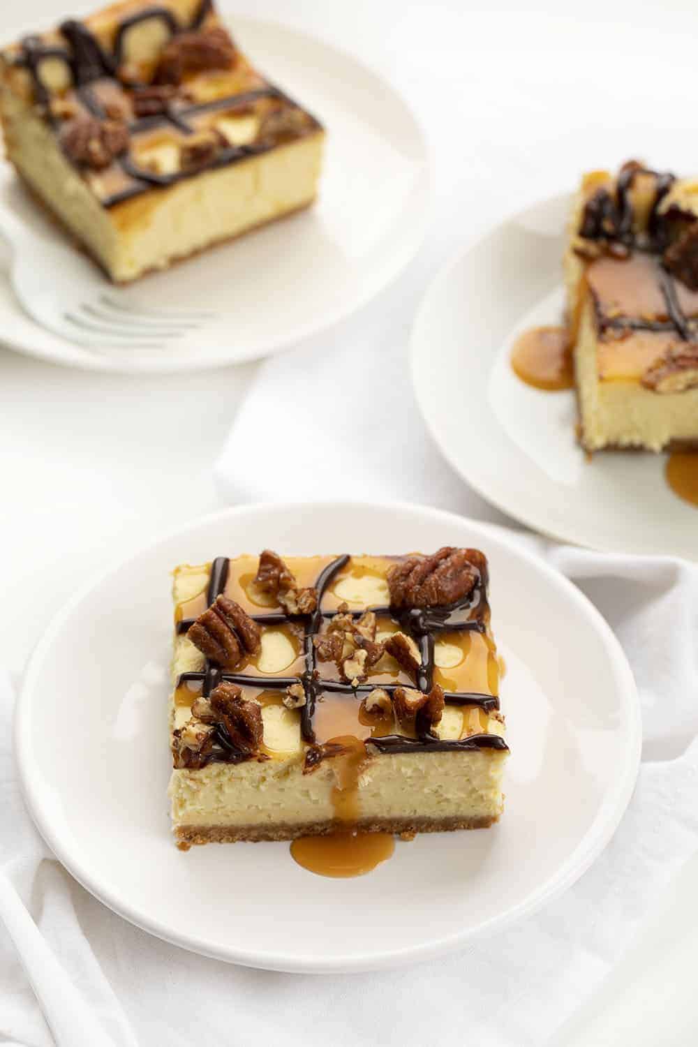 Turtle Cheesecake Bars are creamy dessert bars with a graham cracker crust and cheesecake filling, topped with caramel, candied pecans, and hot fudge. Turtle Cheesecake, Cheesecake Bars, Turtle Cheesecake Bars, Dessert, Cheesecake, Best Cheesecake Recipe, i am baker, iambaker, recipes, baking Pieces of Turtle Cheesecake Bars on Plates