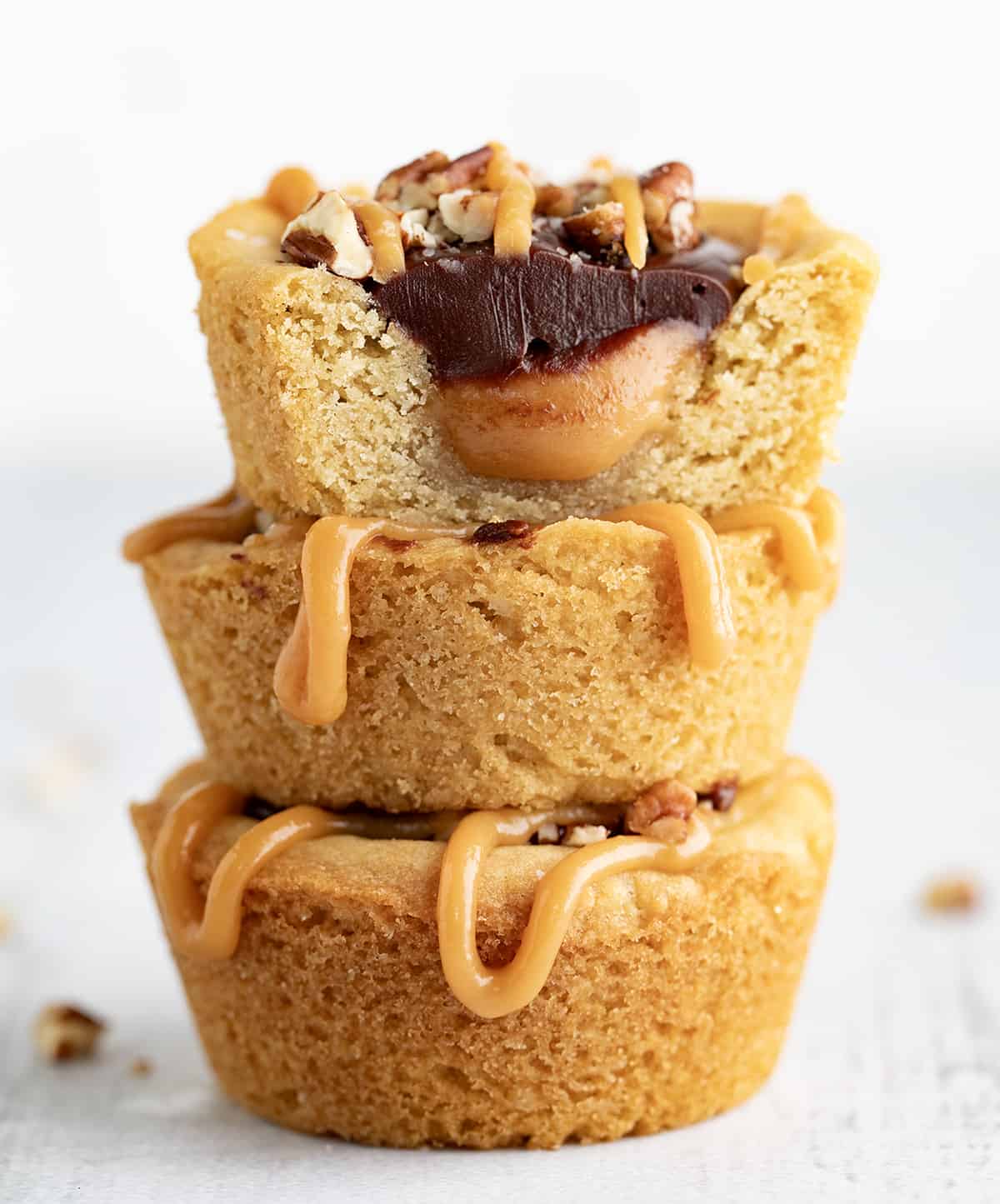 Turtle Cookie Cups are a heavenly combination of soft and chewy cookies, homemade caramel, rich chocolate ganache, crunchy pecans, and a hint of salt. They are a delightfully indulgent treat made with the classic flavors of a turtle dessert in a cookie cup that is baked in a muffin tin. Stacked Turtle Cookie Cups with the Top Cup Cut in Half Showing Inside.