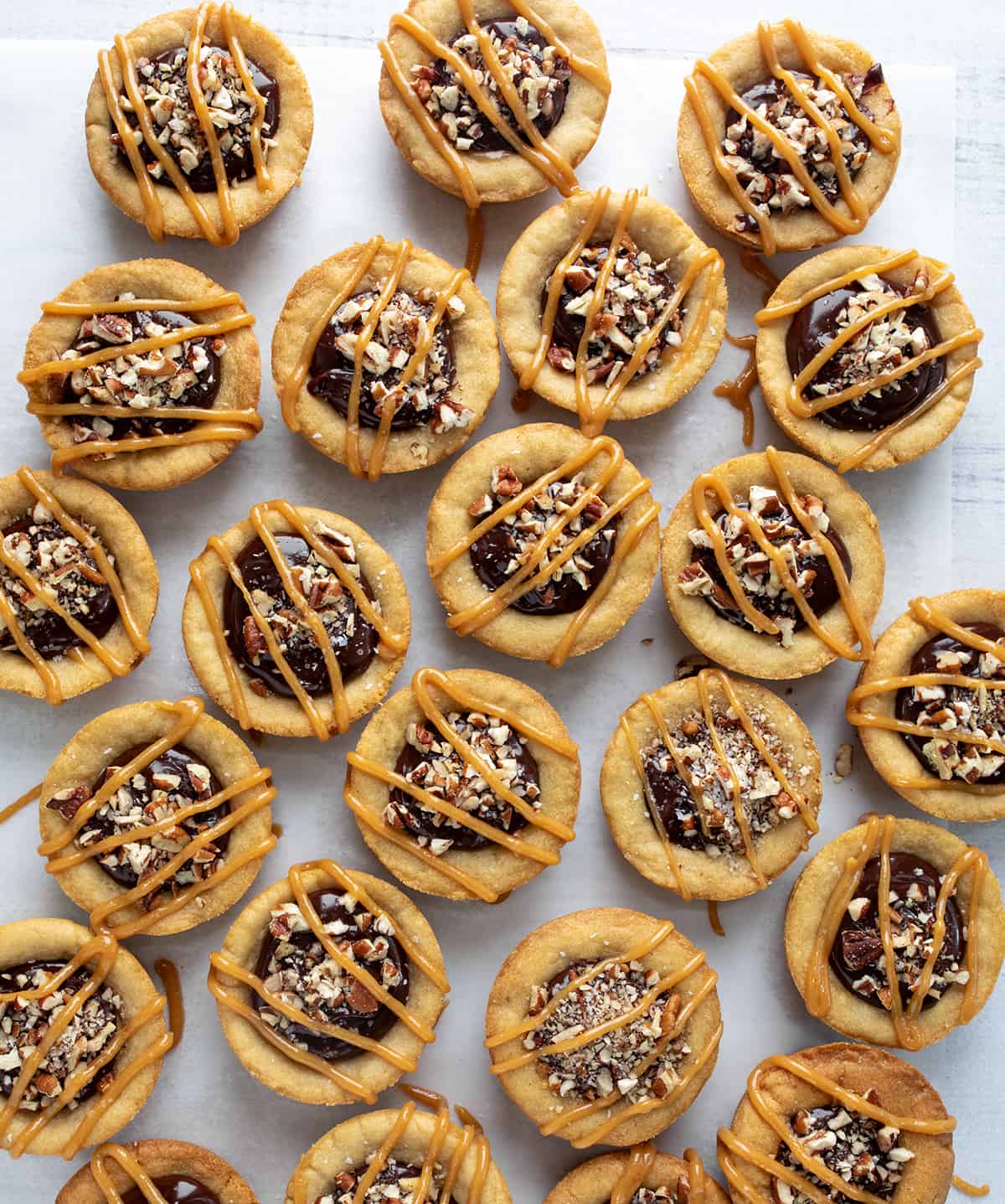 Turtle Cookie Cups are a heavenly combination of soft and chewy cookies, homemade caramel, rich chocolate ganache, crunchy pecans, and a hint of salt. They are a delightfully indulgent treat made with the classic flavors of a turtle dessert in a cookie cup that is baked in a muffin tin. Several Turtle Cookie Cups on a White Counter from Overhead.