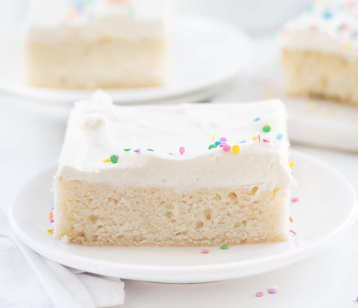 Vanilla Delight Cake (or white cake) is a moist, delicate, and flavorful white sheet cake that is delicious on its own or perfect for your favorite frostings and toppings. It is definitely delightful to snack on, enjoy as a dessert, or share at your next celebration or party! Piece of Vanilla-Delight Cake or White Cake on a White Plate with More Plates in the Background.