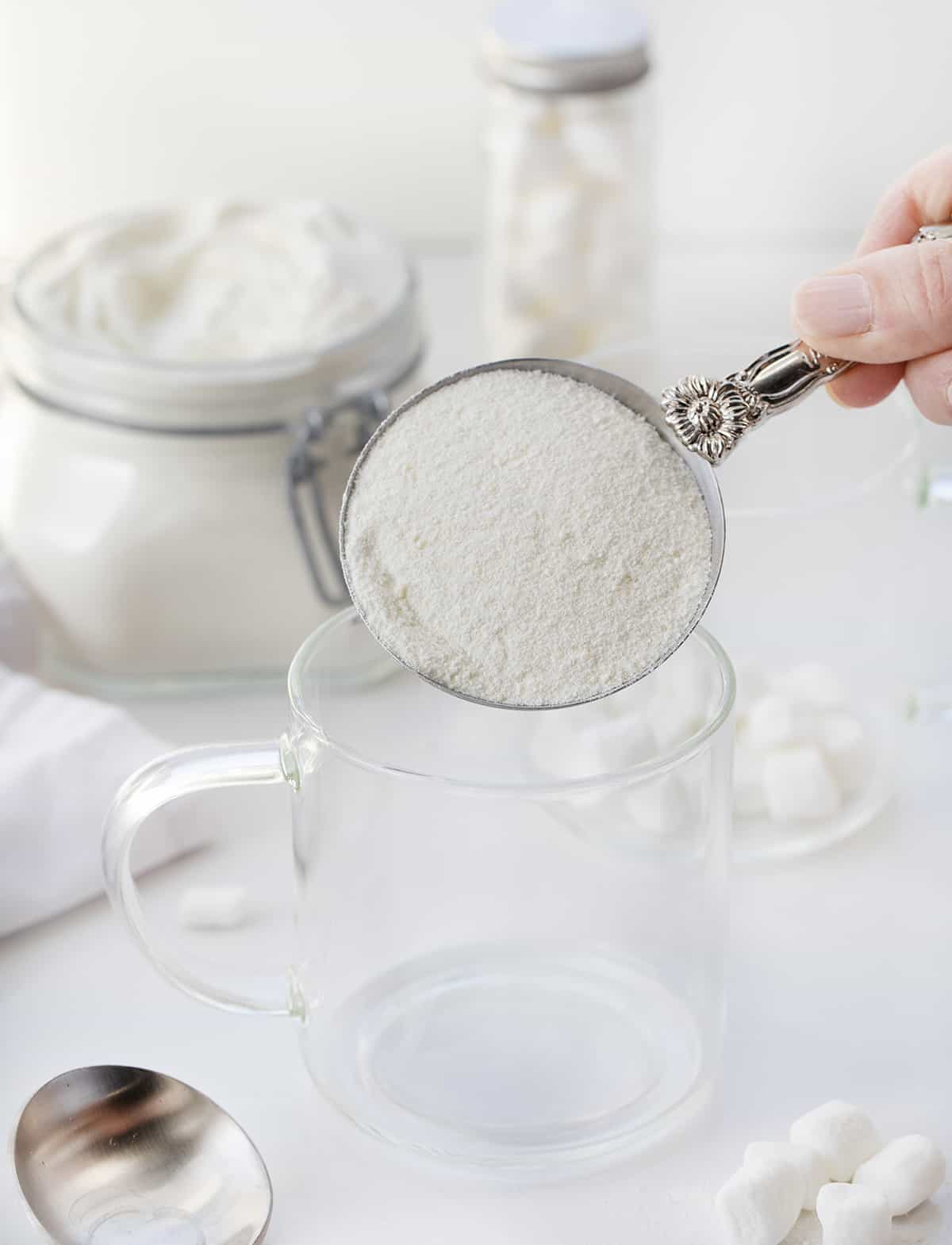 This Big Batch (it makes 20+ cups) of White Hot Chocolate Mix is my new favorite way to enjoy cocoa! As soon as you see how easy it is to make, you will never reach for the packets at the store. It can be added to hot water, hot milk, or even to your cup of coffee. Crockpot Hot Cocoa directions are in the post, which would make it perfect for a Christmas Party, New years Even drink, a mocktail, or a cocktail if you add liquor! And, better yet, it will last up to a year when stored properly. Measuring cup with white chocolate cocoa powder in it about to be pouring in a glass.