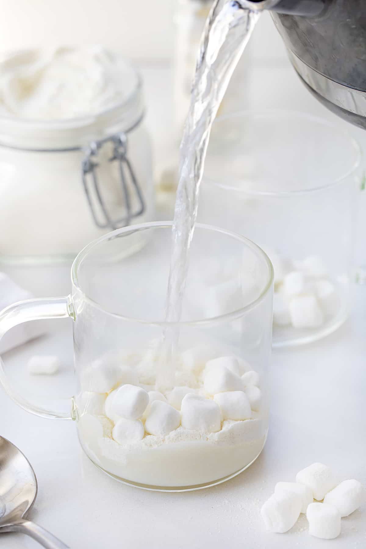 This Big Batch (it makes 20+ cups) of White Hot Chocolate Mix is my new favorite way to enjoy cocoa! As soon as you see how easy it is to make, you will never reach for the packets at the store. It can be added to hot water, hot milk, or even to your cup of coffee. Crockpot Hot Cocoa directions are in the post, which would make it perfect for a Christmas Party, New years Even drink, a mocktail, or a cocktail if you add liquor! And, better yet, it will last up to a year when stored properly. Pouring Hot water over white chocolate hot cocoa mix and mini marshmallows in a clear glass.