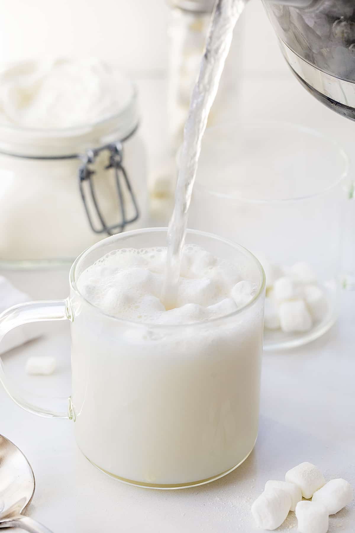This Big Batch (it makes 20+ cups) of White Hot Chocolate Mix is my new favorite way to enjoy cocoa! As soon as you see how easy it is to make, you will never reach for the packets at the store. It can be added to hot water, hot milk, or even to your cup of coffee. Crockpot Hot Cocoa directions are in the post, which would make it perfect for a Christmas Party, New years Even drink, a mocktail, or a cocktail if you add liquor! And, better yet, it will last up to a year when stored properly. Pouring Hot water over white chocolate hot cocoa mix and mini marshmallows in a clear glass.