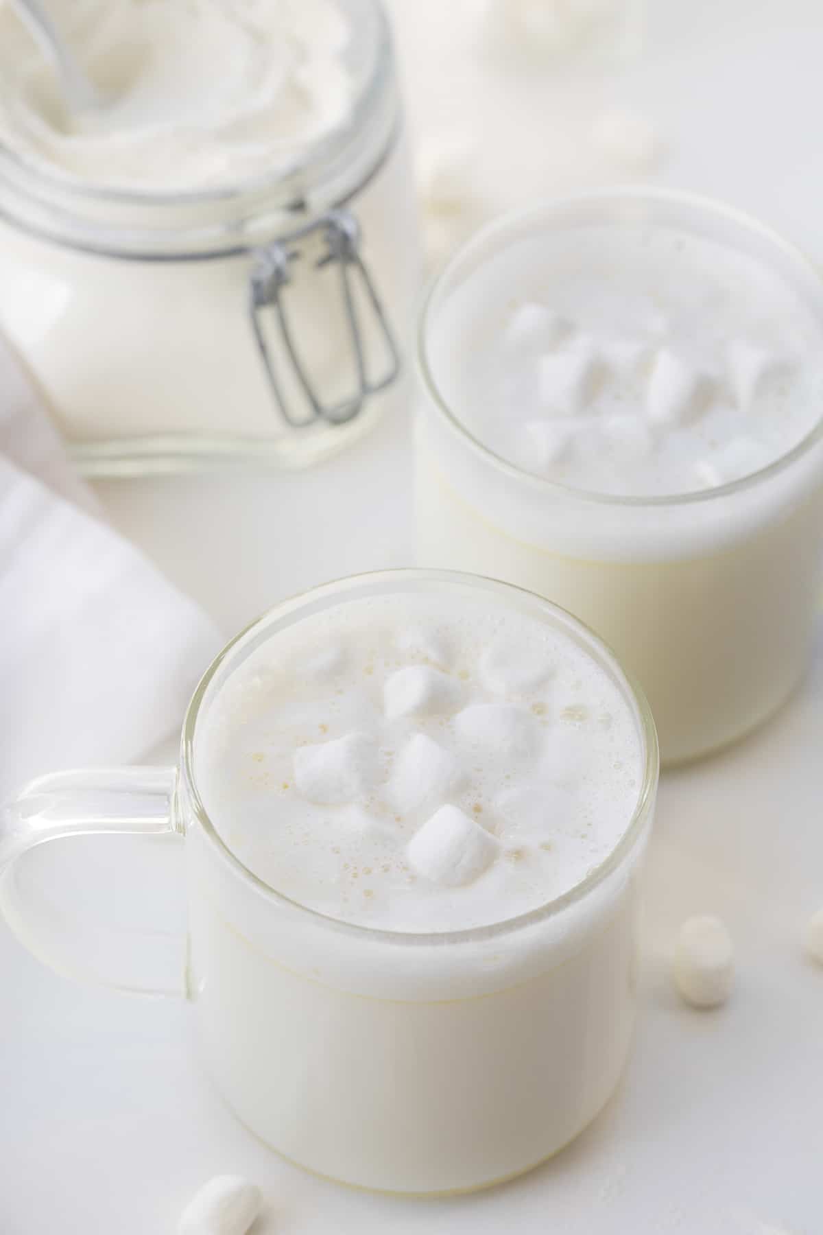 This Big Batch (it makes 20+ cups) of White Hot Chocolate Mix is my new favorite way to enjoy cocoa! As soon as you see how easy it is to make, you will never reach for the packets at the store. It can be added to hot water, hot milk, or even to your cup of coffee. Crockpot Hot Cocoa directions are in the post, which would make it perfect for a Christmas Party, New years Even drink, a mocktail, or a cocktail if you add liquor! And, better yet, it will last up to a year when stored properly. Cups of white hot chocolate on a white counter with mini marshmallows and the cocoa in the background.