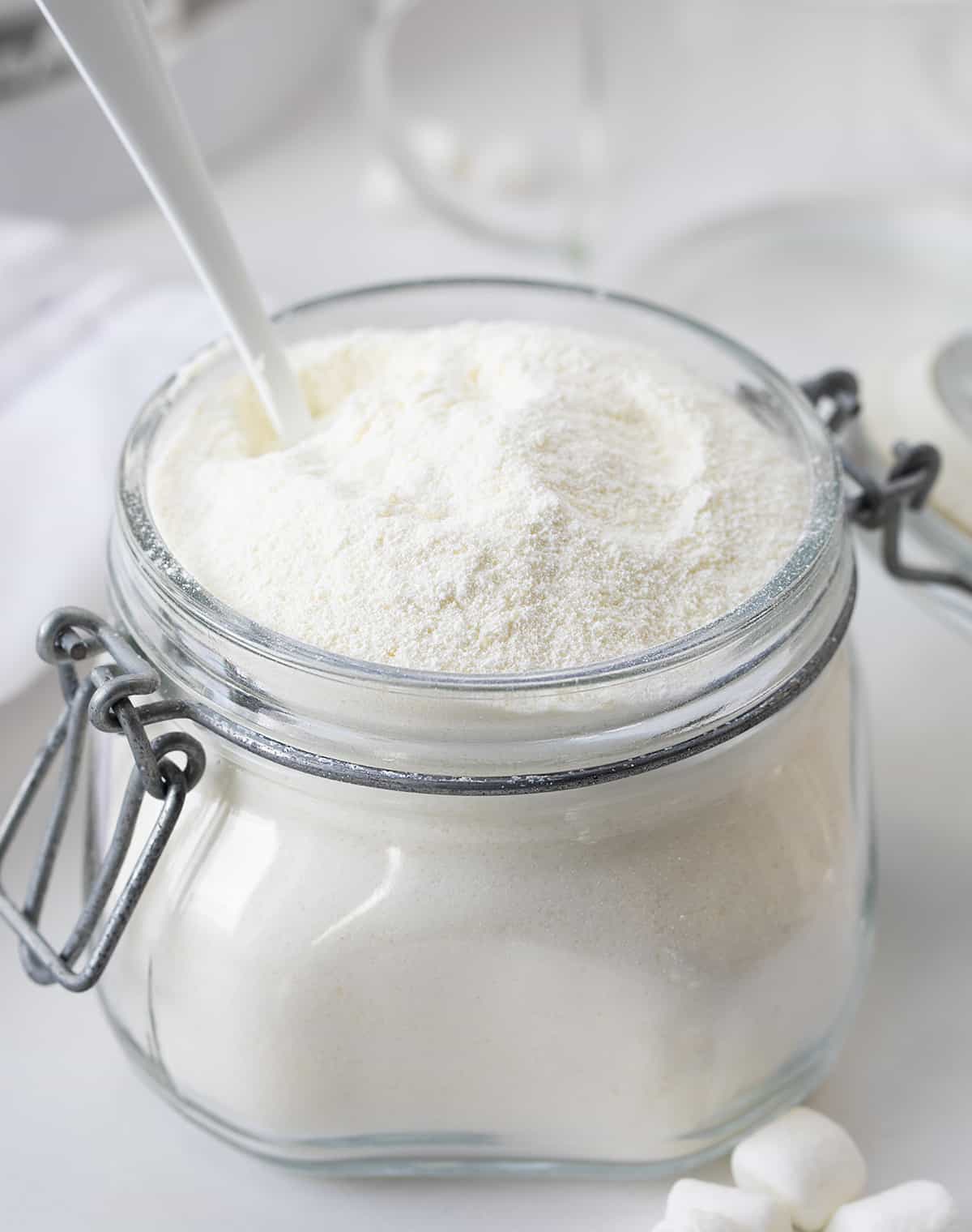 This Big Batch (it makes 20+ cups) of White Hot Chocolate Mix is my new favorite way to enjoy cocoa! As soon as you see how easy it is to make, you will never reach for the packets at the store. It can be added to hot water, hot milk, or even to your cup of coffee. Crockpot Hot Cocoa directions are in the post, which would make it perfect for a Christmas Party, New years Even drink, a mocktail, or a cocktail if you add liquor! And, better yet, it will last up to a year when stored properly. Container filled with homemade white hot chocolate powder and a spoon in it.