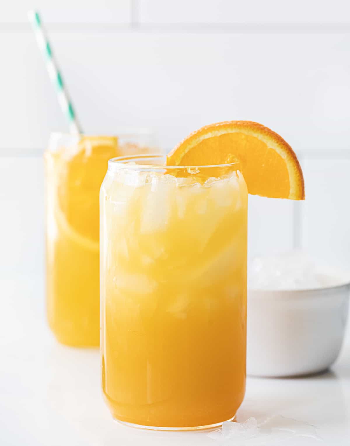 Yellow Hammer Slammer is an adult cocktail made with vodka, pineapple rum, amaretto, orange juice, and pineapple juice. It's a refreshing drink that is based on a cocktail that was created in Gallettes bar in Tuscaloosa, Alabama. It's their signature drink (Yellow Hammer) that is most popular on game day, meaning Alabama football games. But, you can recreate the drink at home, no need to wait for football season! Glasses of Yellow Hammer Slammer with an Orange Garnish on a White Counter.